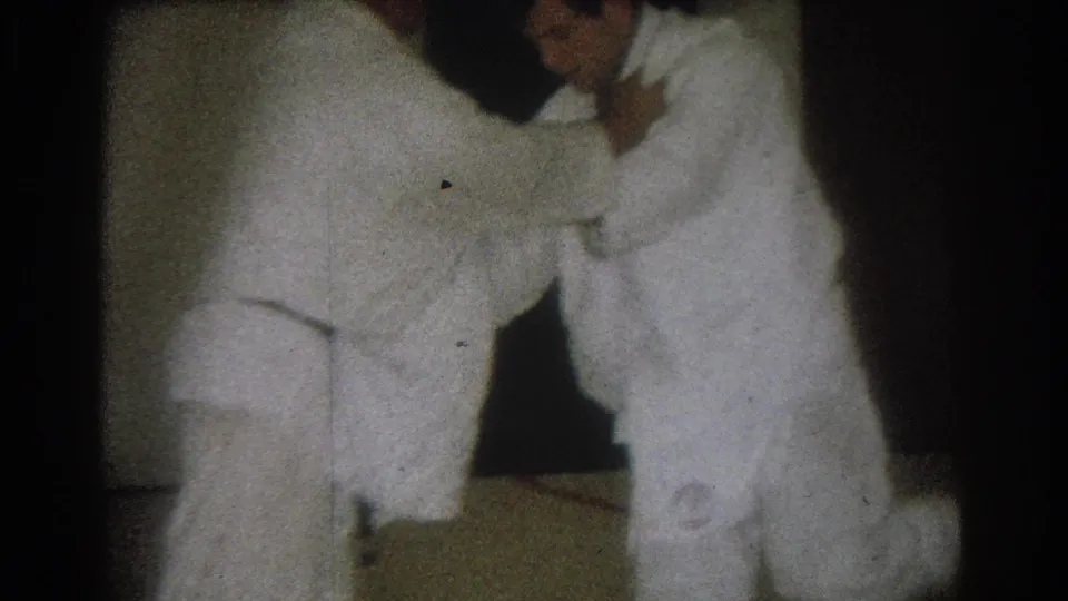 Two men in white gi continue a standing martial arts hold in a dim indoor space in the West Covina 1966 clip.