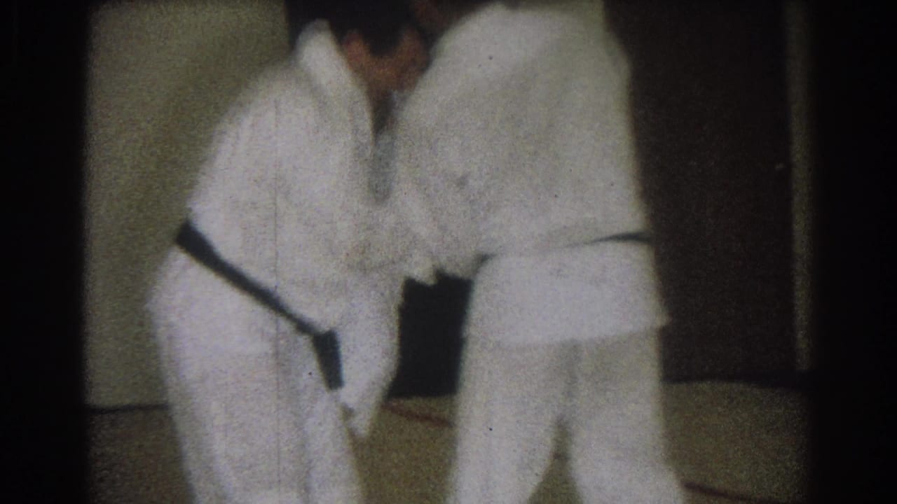 Two black-belted adult men in white gis lean forward during a judo practice move in a dim private room.