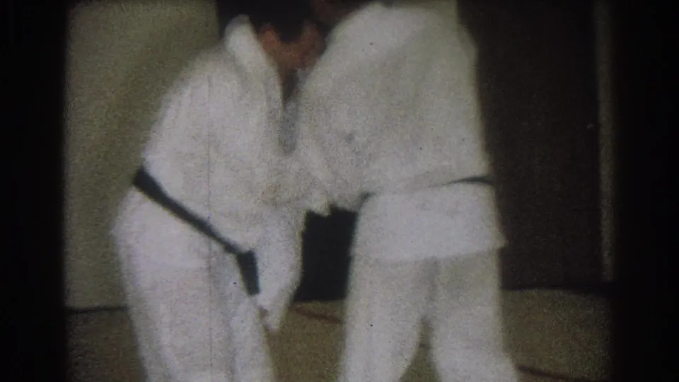Two black-belted adult men in white gis lean forward during a judo practice move in a dim private room.