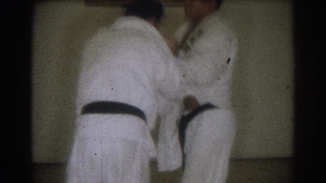 Two adult men in white gis continue a close judo drill inside a dim room, captured with grain and soft motion blur.