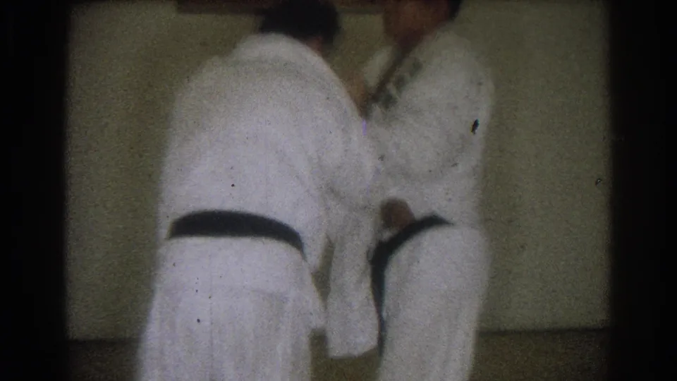 Two adult men in white gis continue a close judo drill inside a dim room, captured with grain and soft motion blur.