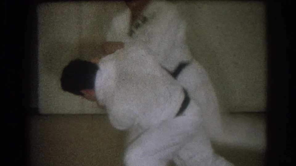In 1966 West Covina home-movie footage, two adult men in white judo gis are locked in a close grappling position on a dojo mat.