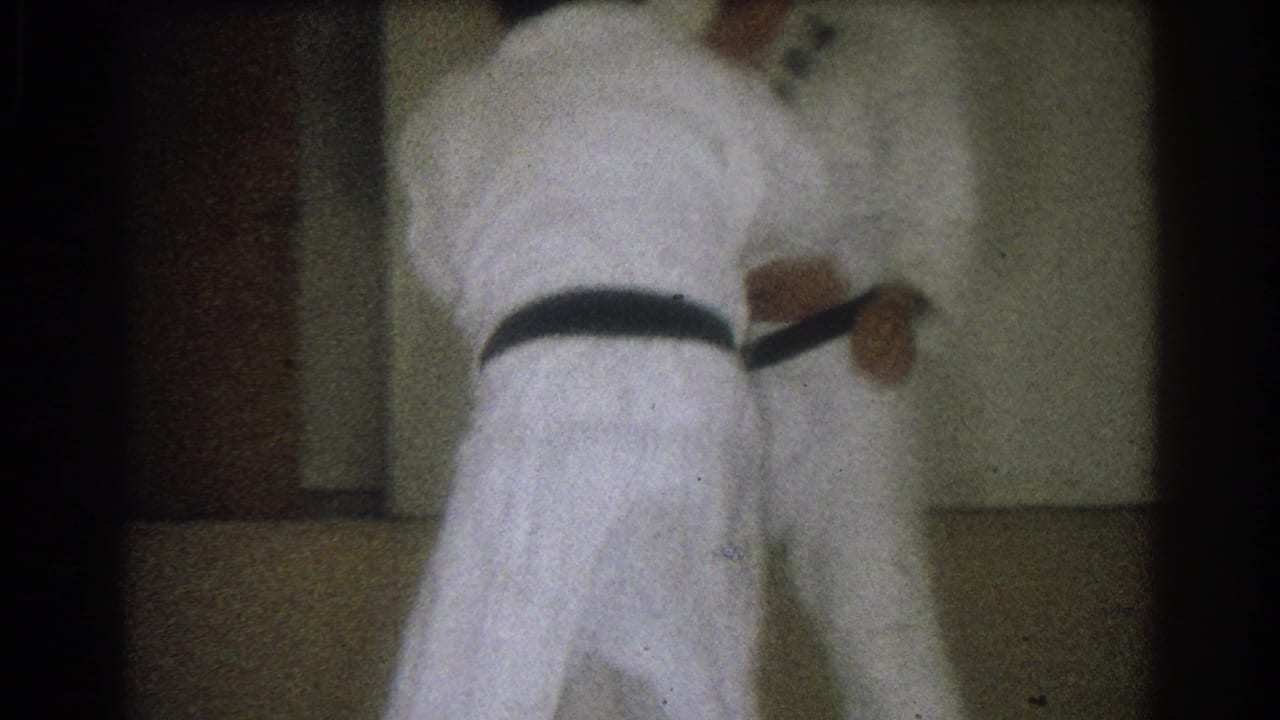 Two adult judo practitioners in white gis stand near the dojo wall, with one black belt clearly visible in the 1966 West Covina footage.