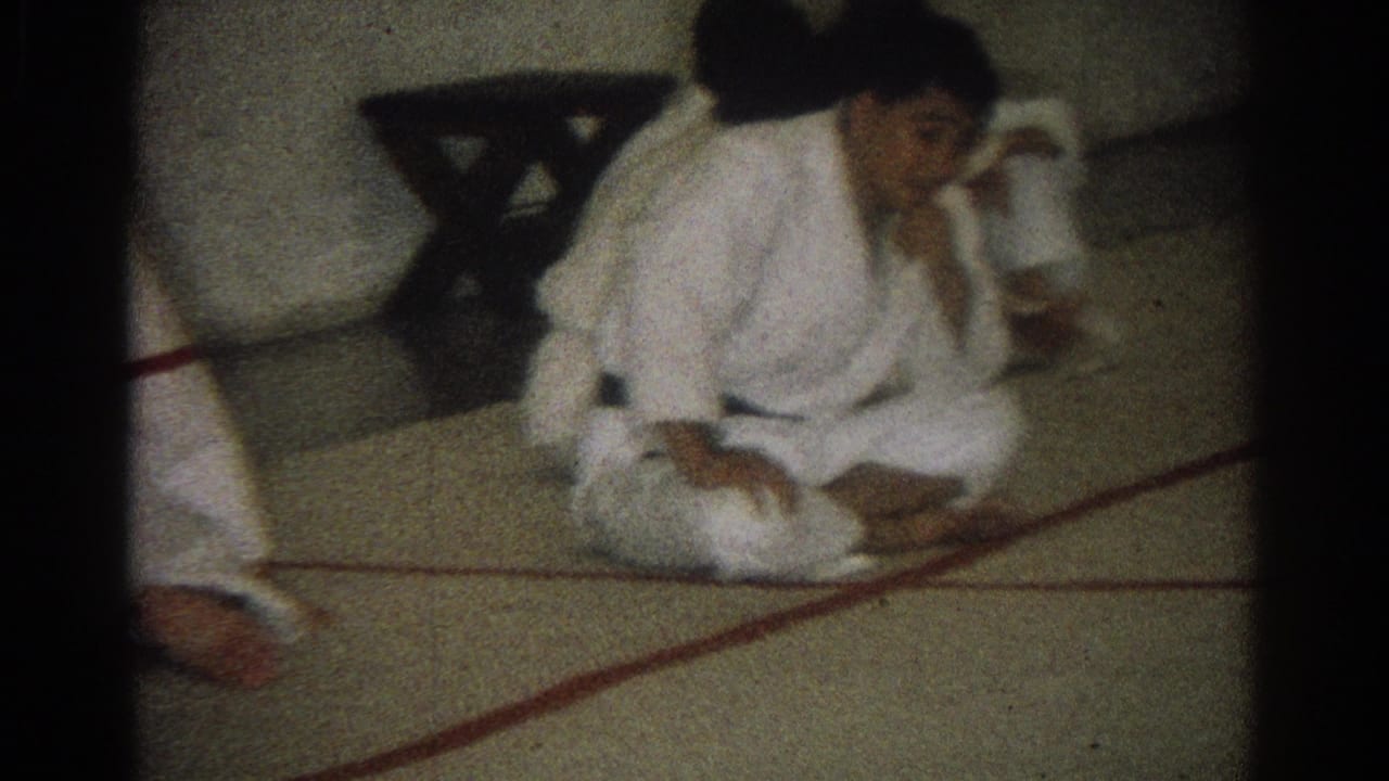 Practitioners in white gi uniforms sit and move on a matted floor with red boundary lines.