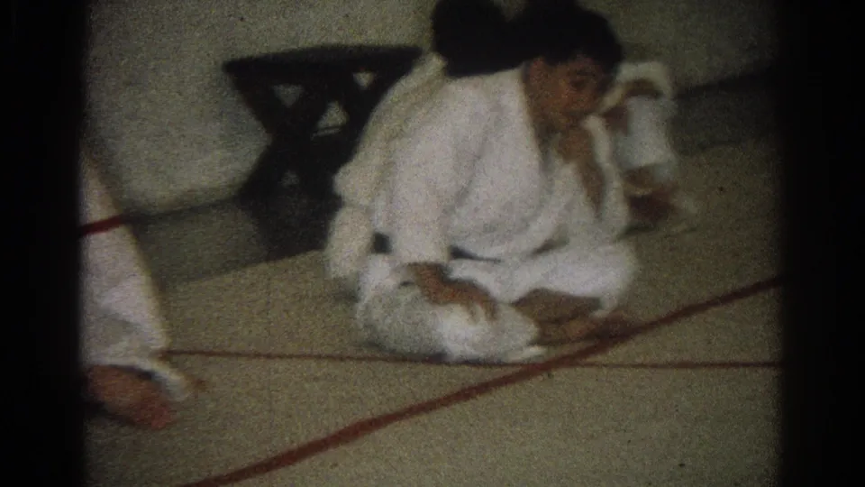 1966 West Covina home-movie frame showing martial arts practitioners in white gi uniforms on an indoor mat, with one seated near red floor lines.