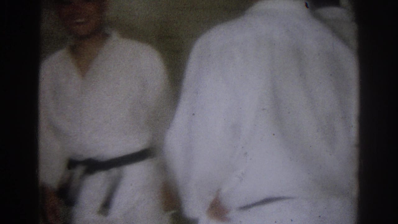 West Covina 1966 frame showing a black belt practitioner in a white gi beside another gi-clad participant during indoor martial arts practice.