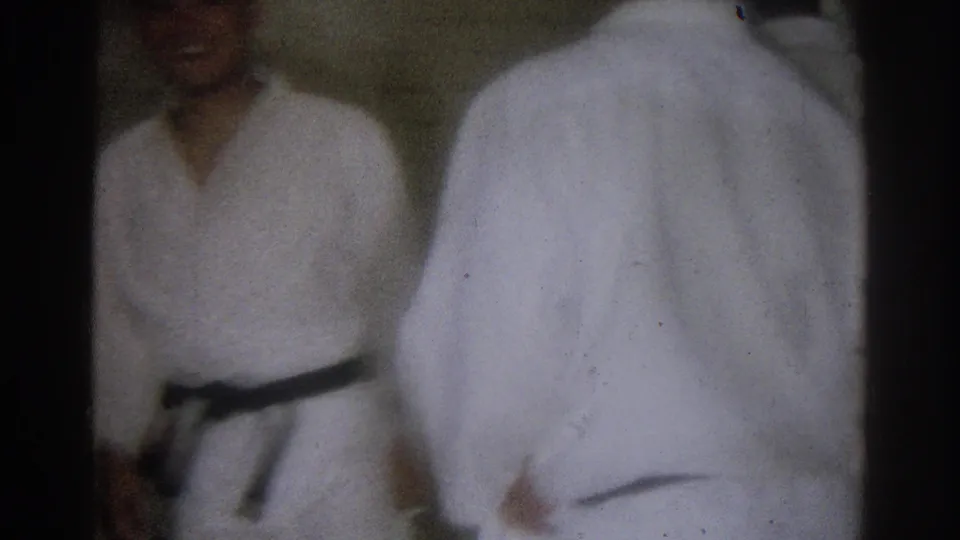 West Covina 1966 frame showing a black belt practitioner in a white gi beside another gi-clad participant during indoor martial arts practice.