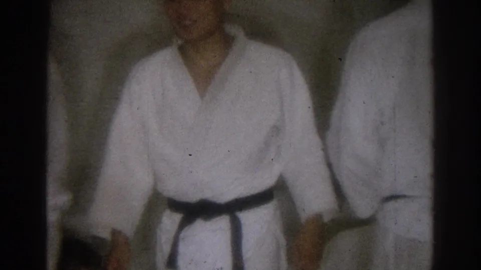 Soft 8mm frame from a 1966 West Covina martial arts session, with a black belt practitioner facing the camera beside another participant.