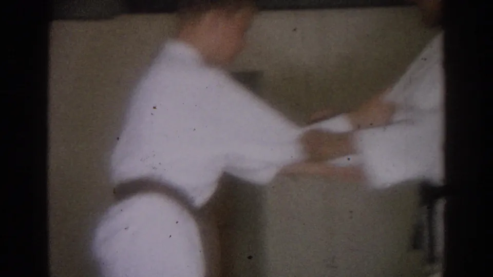 In 1966 West Covina footage, two men in white gi appear to practice a karate attack or block inside a dim room.