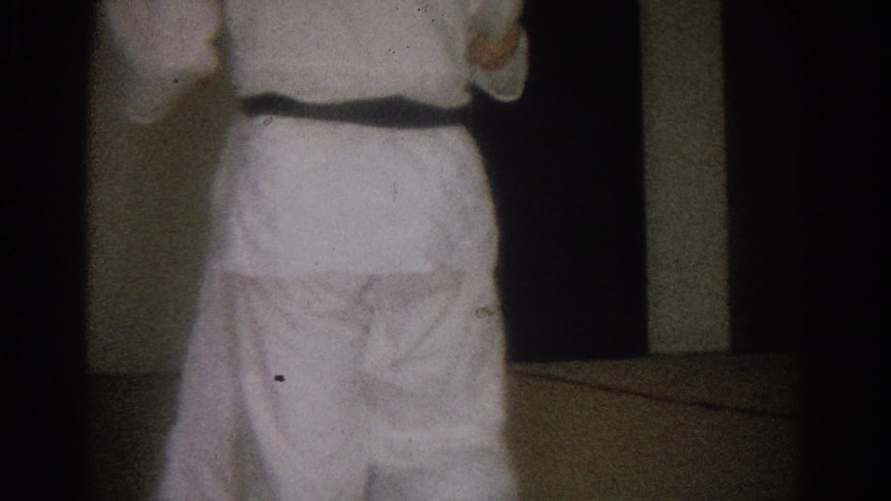 A man in a white karate uniform, black belt visible, stands with his back to the camera in the West Covina home-movie frame.