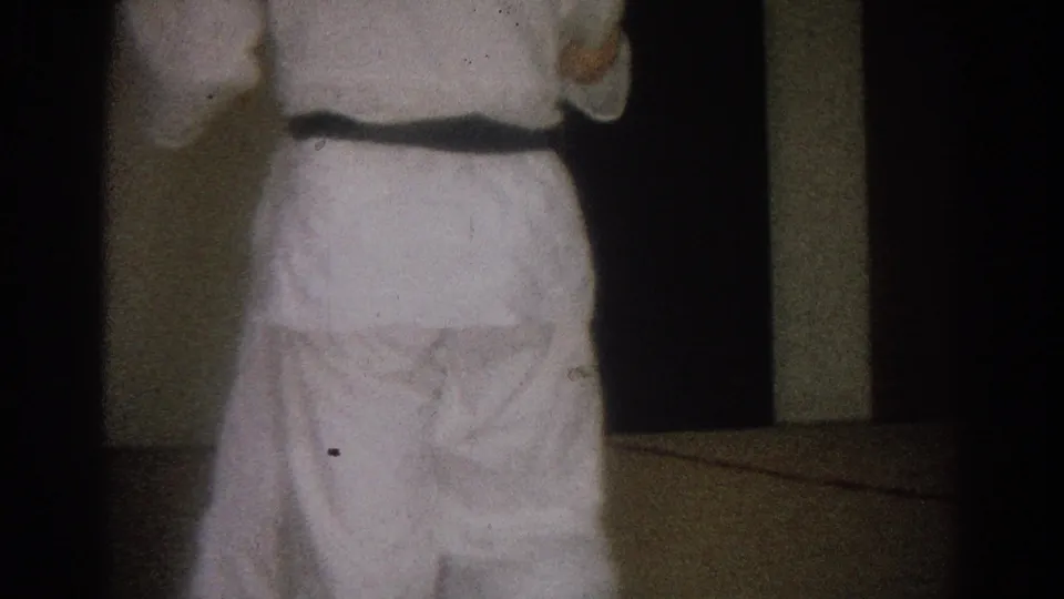 A man in a white karate uniform, black belt visible, stands with his back to the camera in the West Covina home-movie frame.
