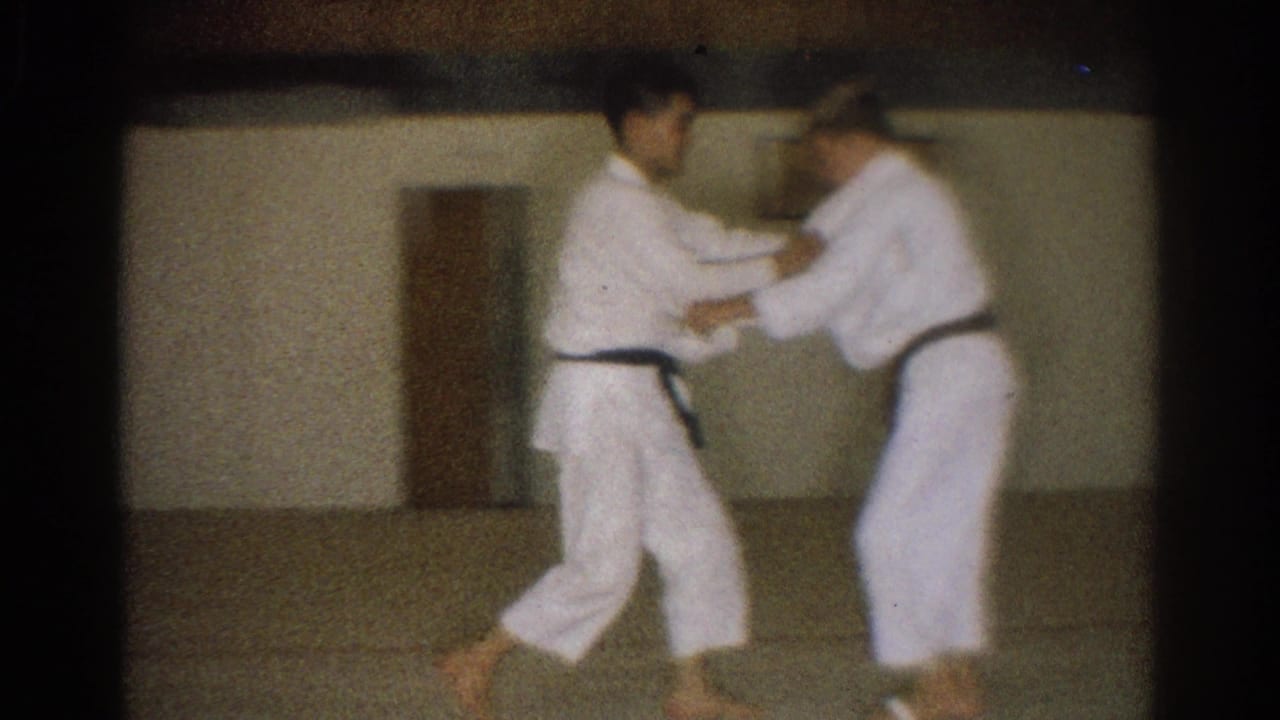 Two young men in white gi and dark belts work through a close sparring hold in the dim West Covina dojo.