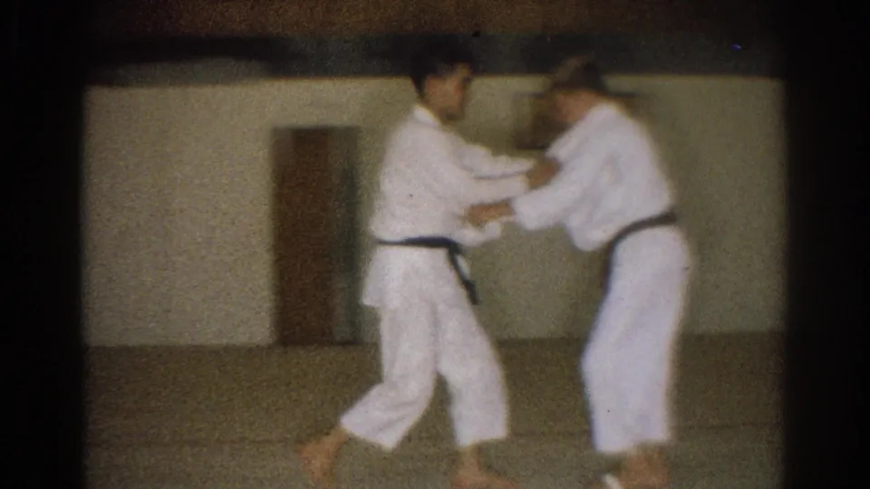Two young men in white gi and dark belts work through a close sparring hold in the dim West Covina dojo.