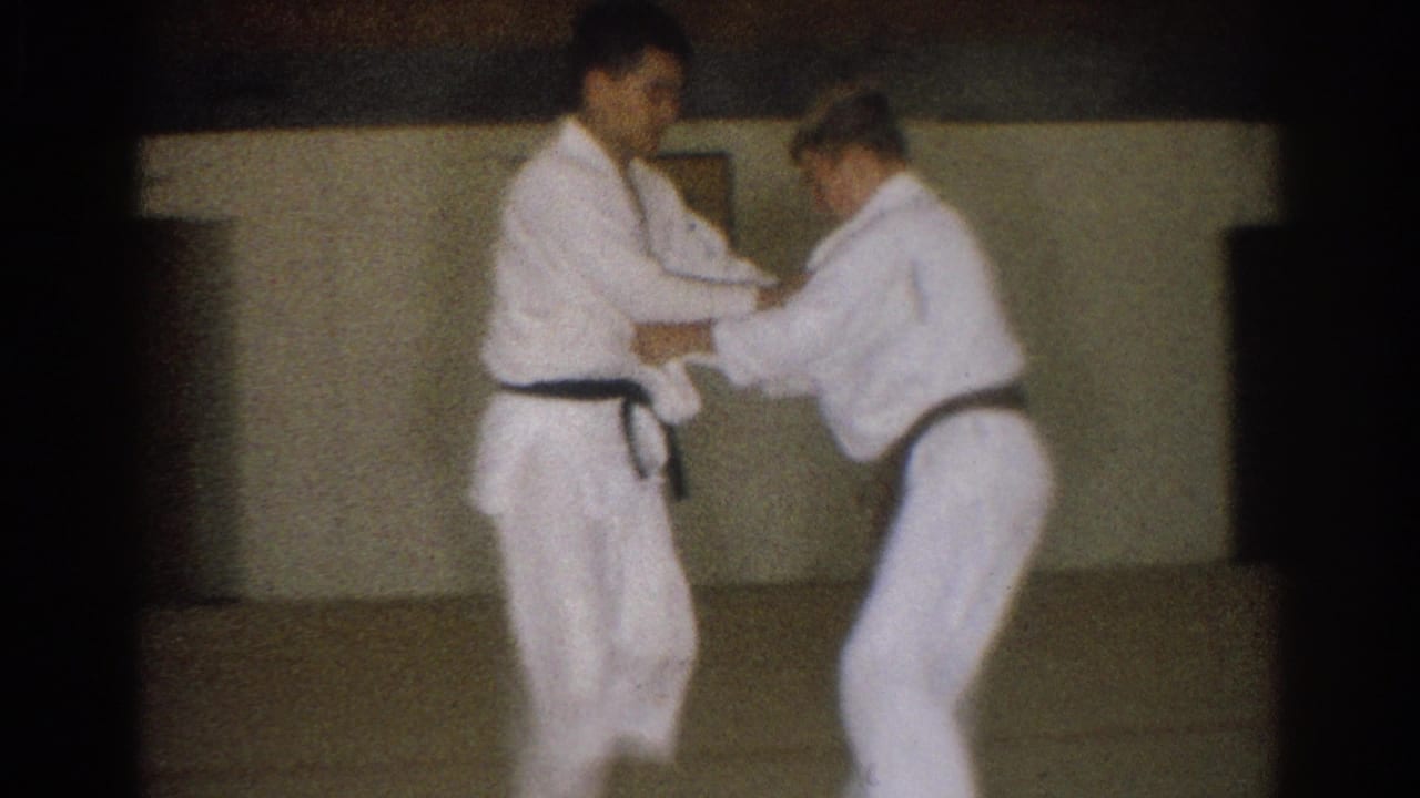 In metadata-supported 1966 West Covina footage, two adult men in white gi and black belts practice a close-contact karate technique indoors.