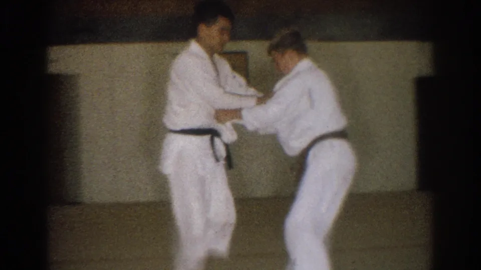In metadata-supported 1966 West Covina footage, two adult men in white gi and black belts practice a close-contact karate technique indoors.