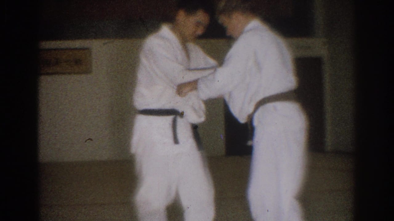 Two men in white gi stand close together while practicing a hand technique indoors.