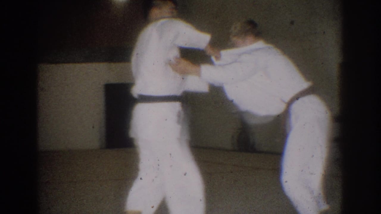 Two judo practitioners in white gis grip each other in a plain indoor room.