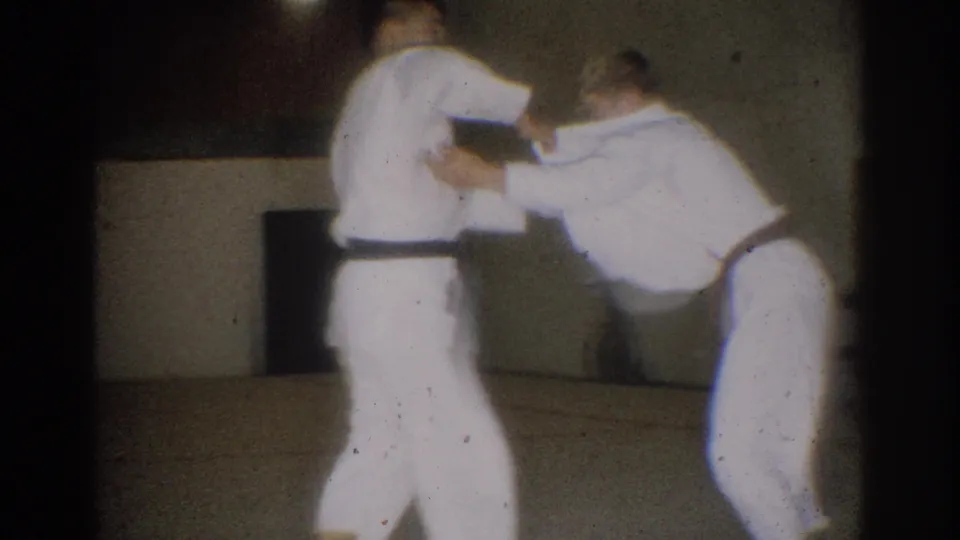 Two young men in white judo gis and dark belts practice a grip sequence in a West Covina indoor dojo, 1966 metadata supplied.