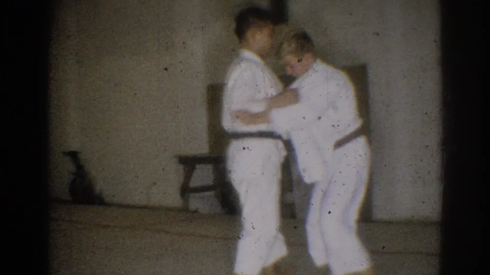 Two black-belt judo practitioners work through a close grappling position inside the simple West Covina training room.