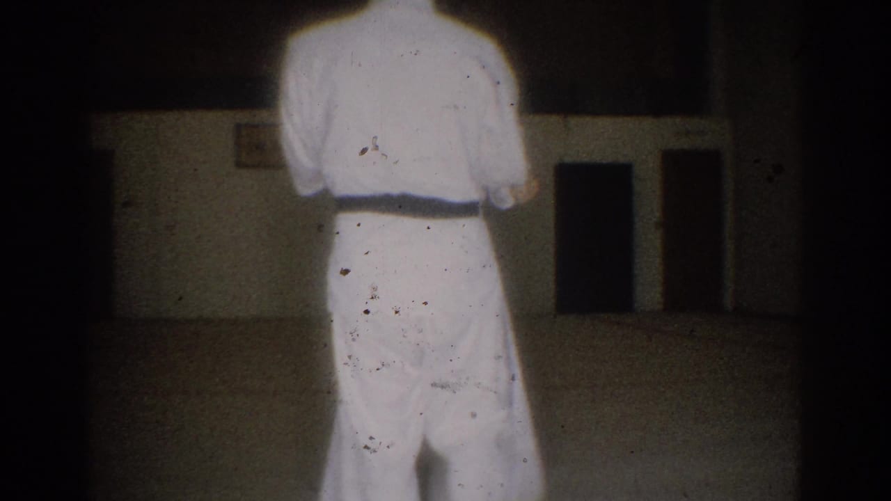 A student in a white gi and dark belt stands from behind in the supplied 1966 West Covina martial arts practice footage.