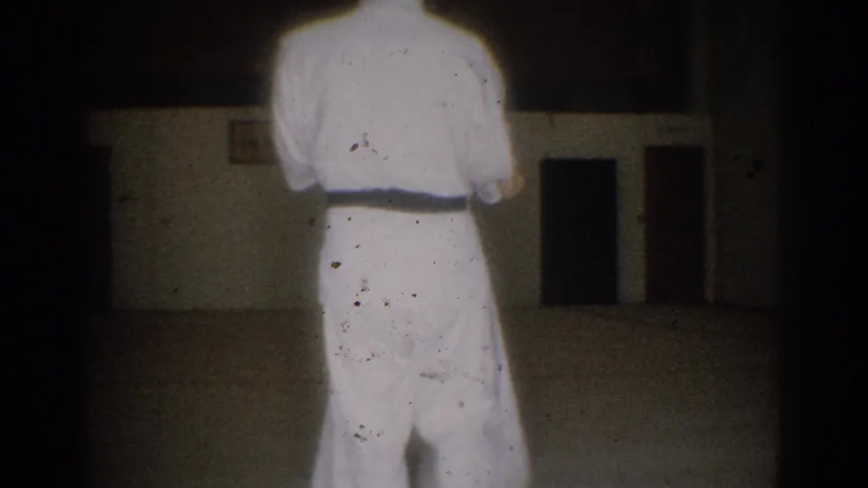 A student in a white gi and dark belt stands from behind in the supplied 1966 West Covina martial arts practice footage.