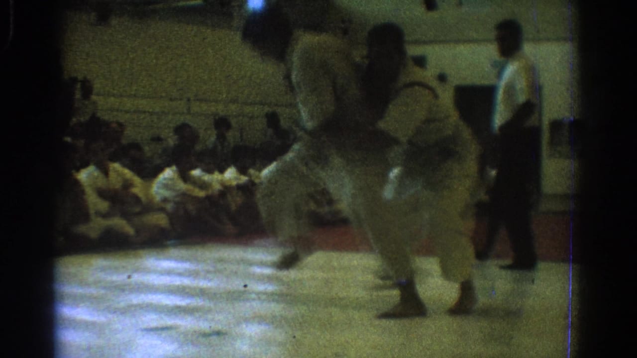 Two martial artists in white gi grapple on a gym mat as spectators sit along the wall.
