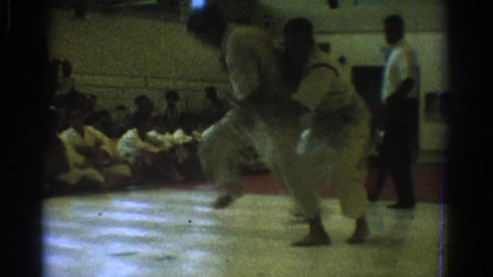 In 1966 West Covina, two martial artists in white gi move through a grappling exchange on a gym mat with seated spectators behind them.