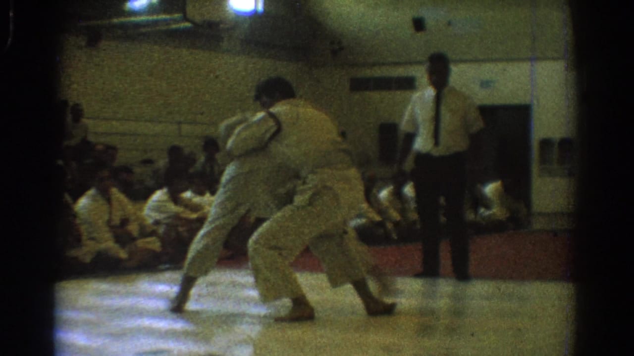 A 1966 West Covina karate match shows two men in white gi crouched in a grappling exchange as a referee observes in the gym.