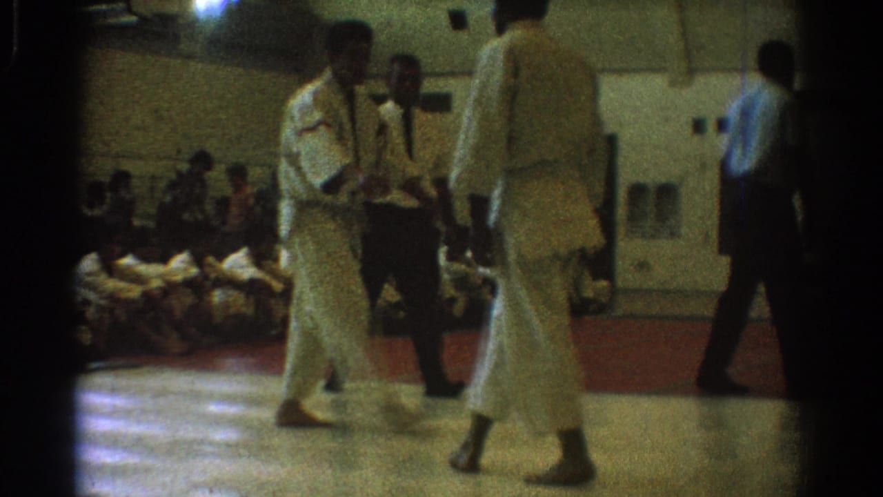 Two uniformed martial artists stand on a gym floor while a referee and spectators watch.