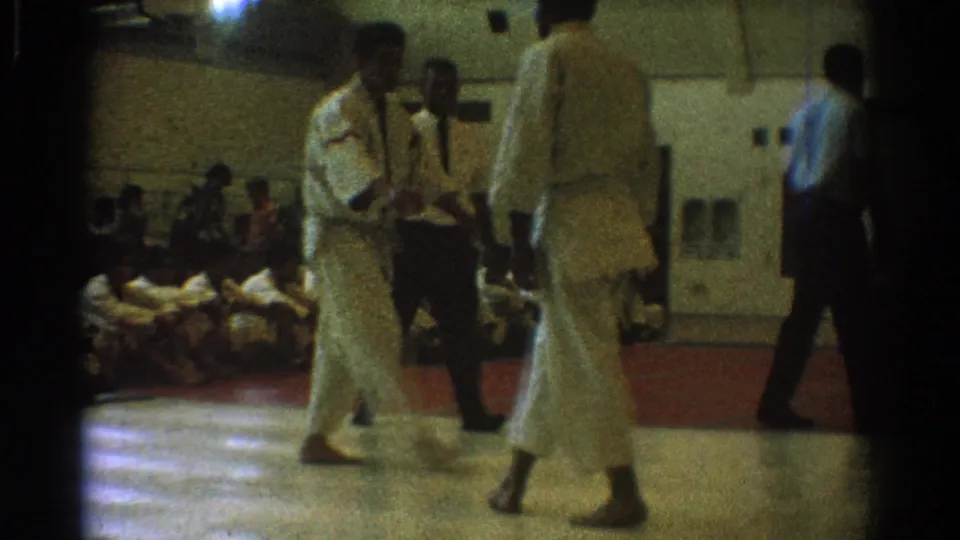 1966 West Covina home-movie frame showing two men in white martial arts uniforms near a referee inside a gymnasium.