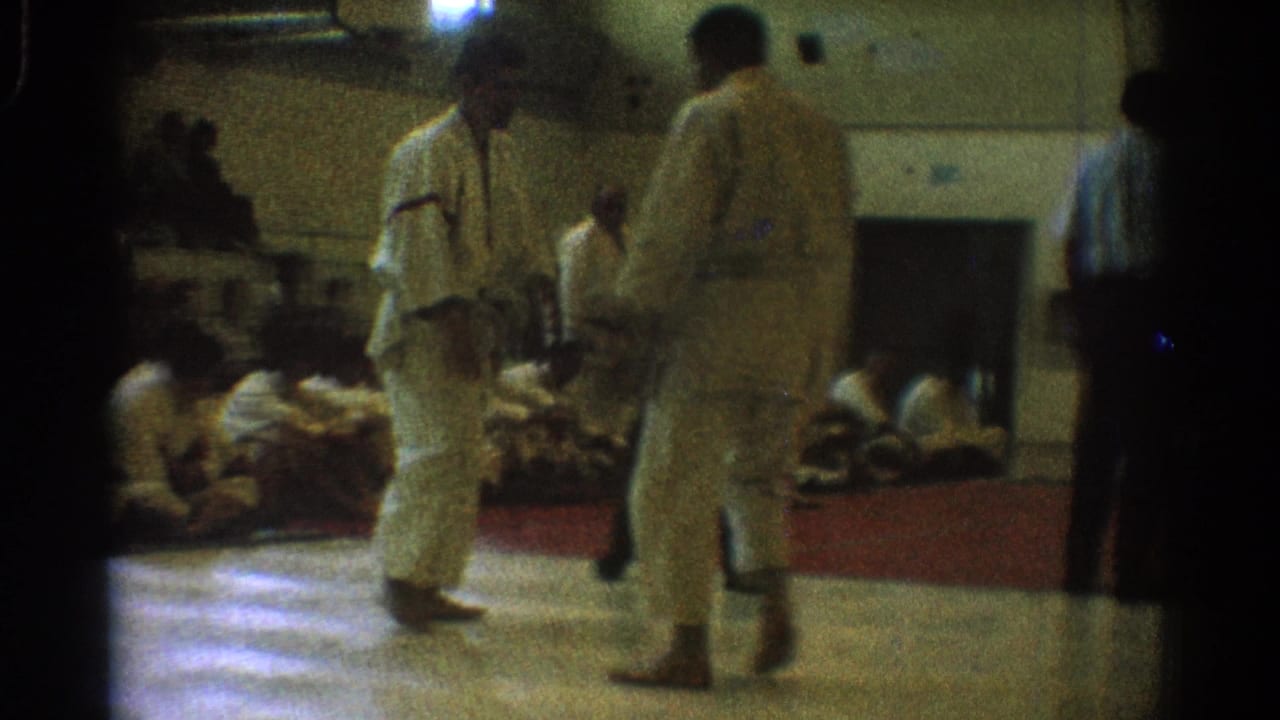 Two uniformed competitors continue a martial arts match in a West Covina gym, with spectators seated along the mat.