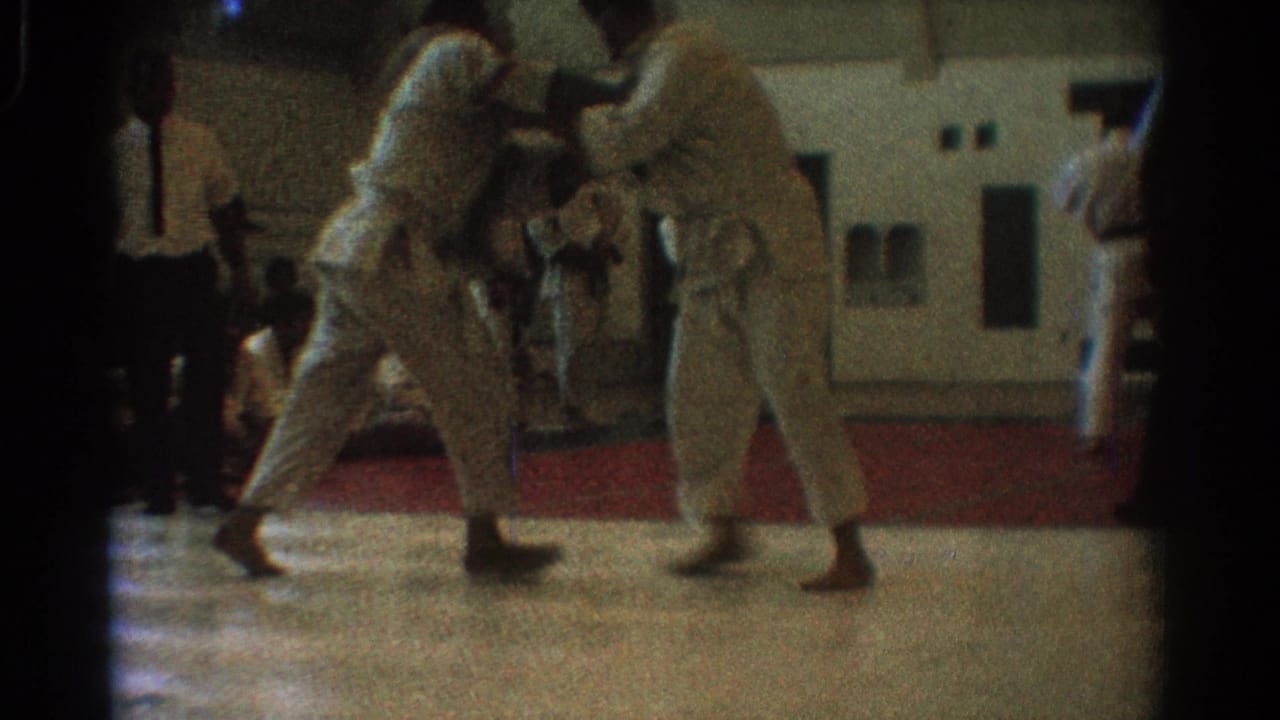 Two martial artists in white uniforms grapple on a gym floor near a red mat edge.