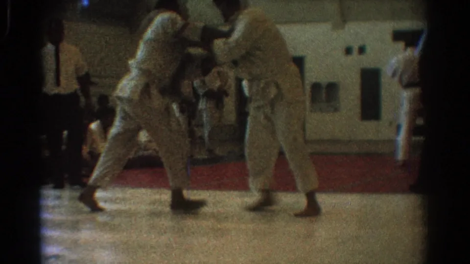 In supported 1966 West Covina home-movie footage, two uniformed martial artists face each other while spectators watch from the gym sidelines.
