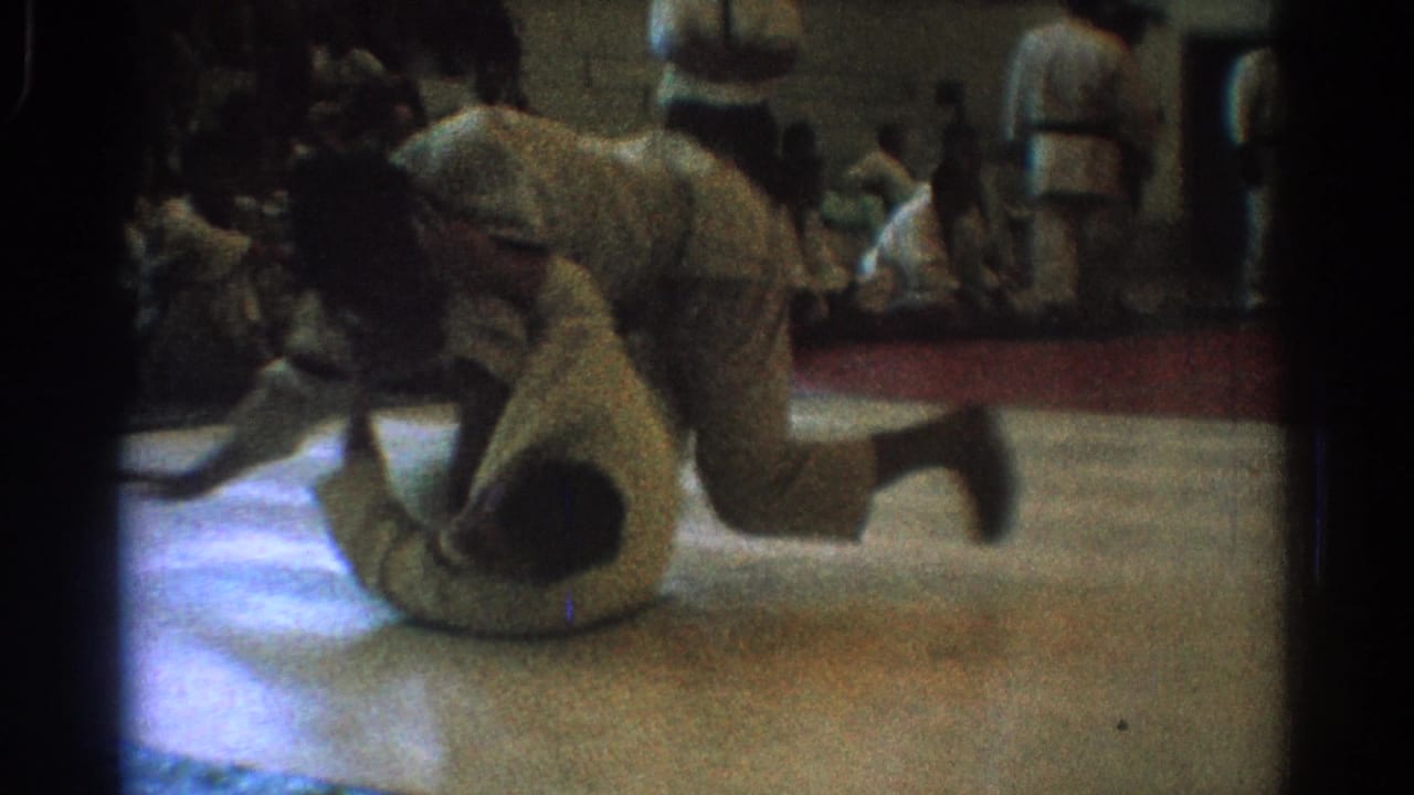 The match continues with one martial artist on the gym floor and another leaning forward, captured in soft, grainy 8mm home-movie framing.
