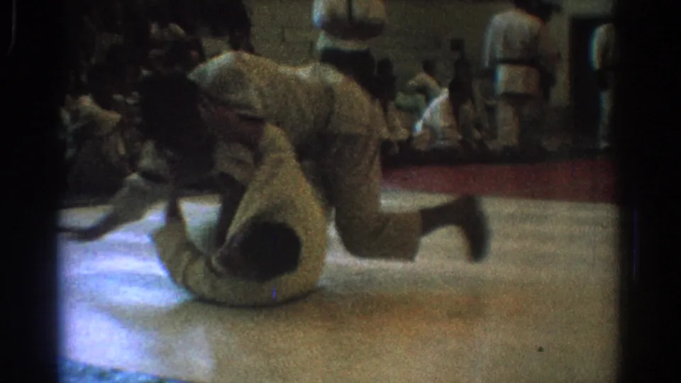 The match continues with one martial artist on the gym floor and another leaning forward, captured in soft, grainy 8mm home-movie framing.