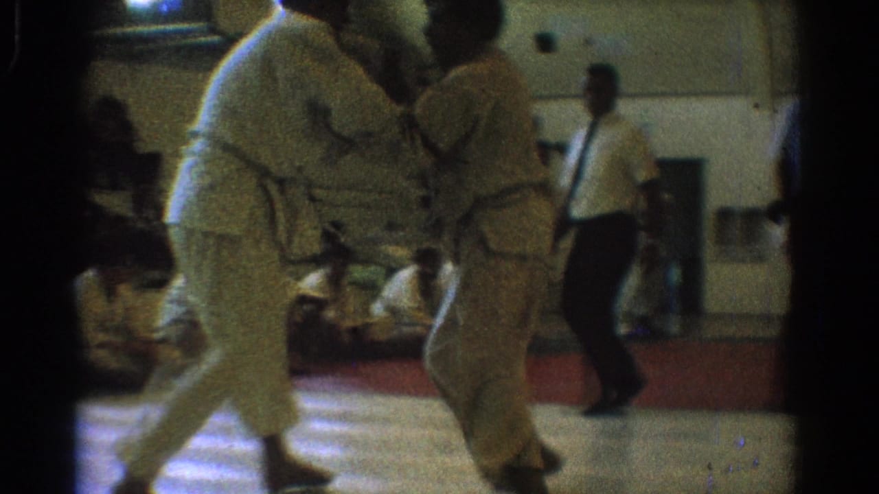 Two uniformed martial artists stand close in the gymnasium match while an official and seated spectators remain visible in the background.