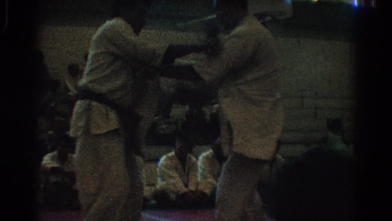 Two men in white uniforms grapple on a red indoor mat with seated spectators behind them.