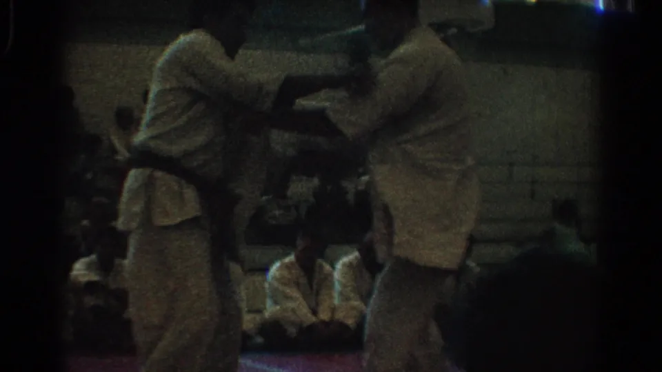 1966 West Covina 8mm frame showing two men in white martial arts uniforms grappling on a red gym mat before seated spectators.