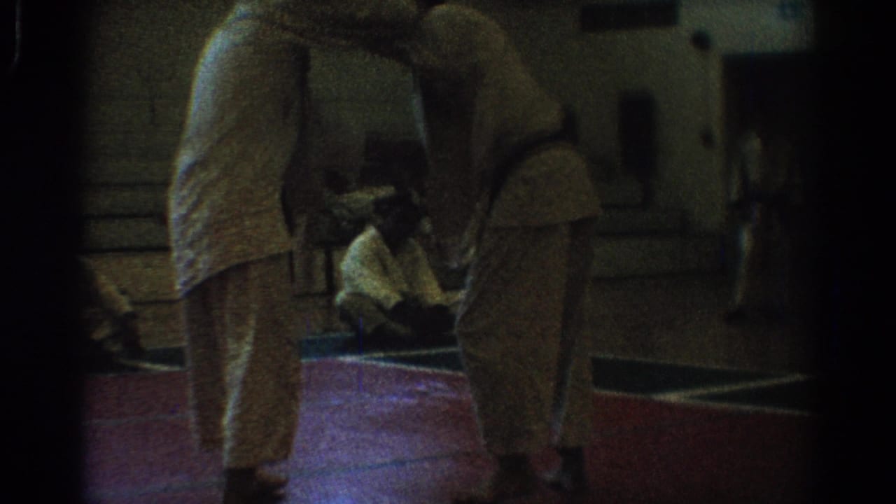 1966 West Covina home-movie frame of two men in white uniforms facing each other on a red indoor mat in a gym.