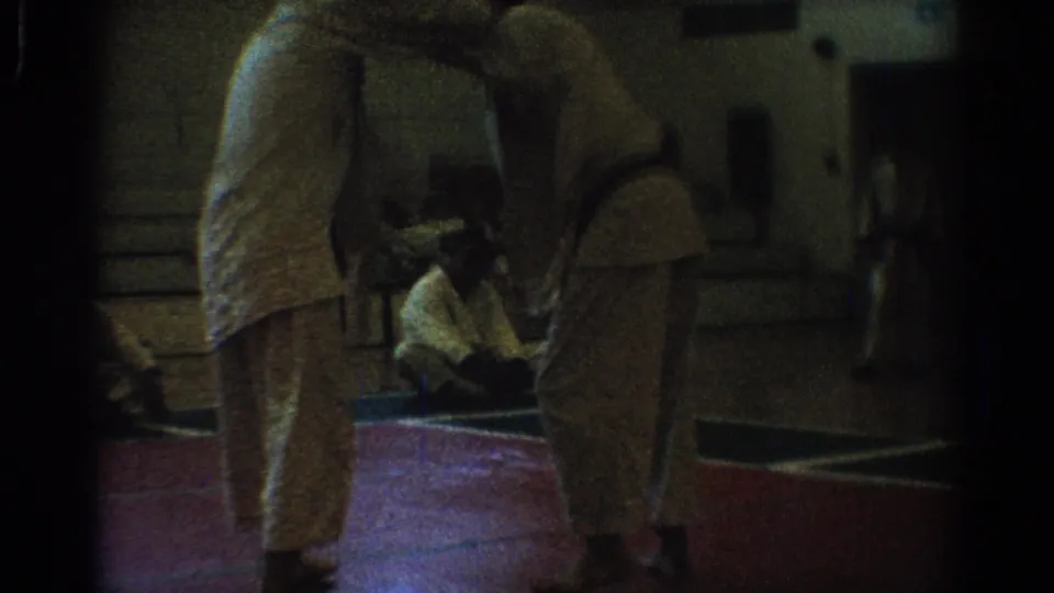 1966 West Covina home-movie frame of two men in white uniforms facing each other on a red indoor mat in a gym.