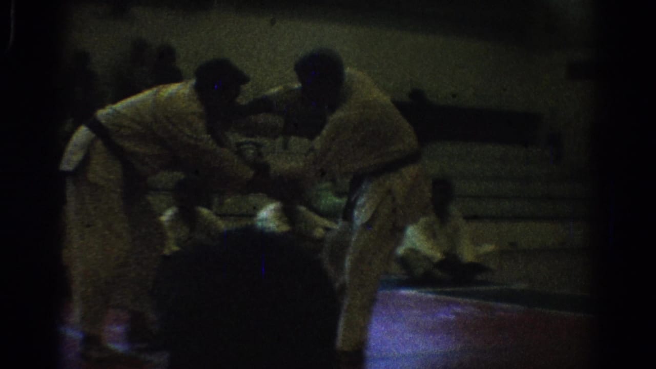Dim 1966 West Covina 8mm frame showing two men in white uniforms leaning into a grappling hold on an indoor mat.