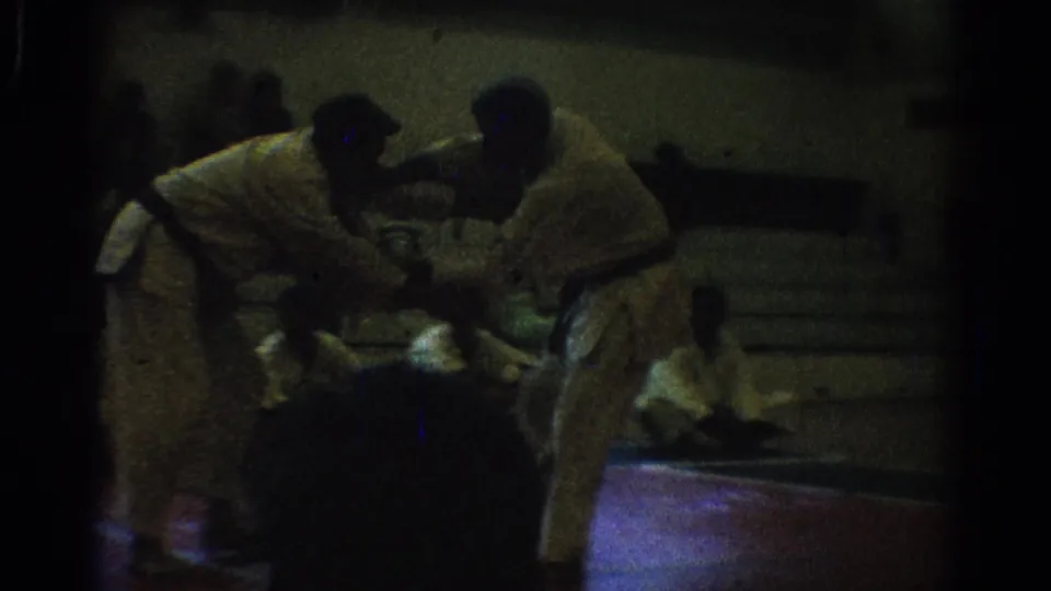 Dim 1966 West Covina 8mm frame showing two men in white uniforms leaning into a grappling hold on an indoor mat.