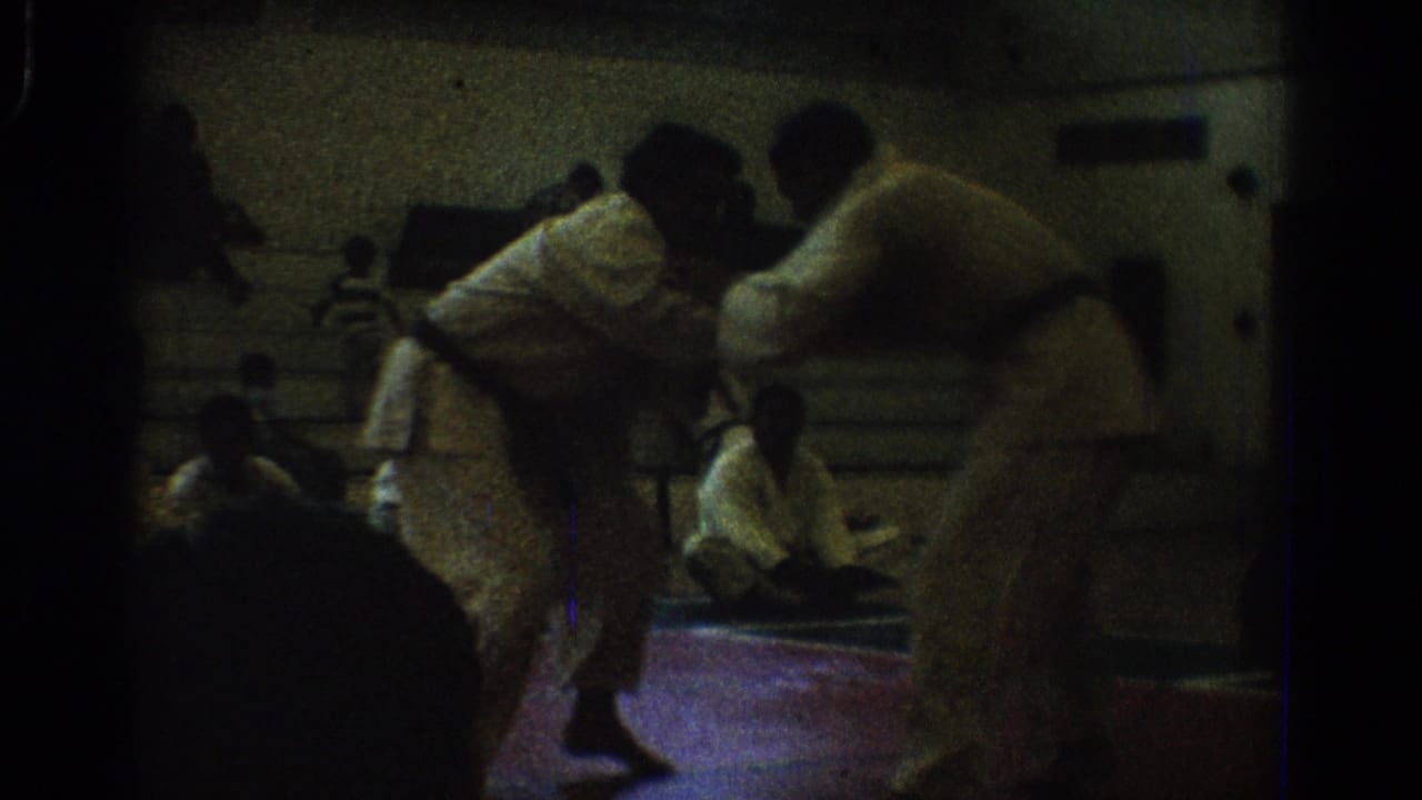1966 West Covina 8mm frame of two uniformed men grappling low on a red indoor mat while people sit along the wall.