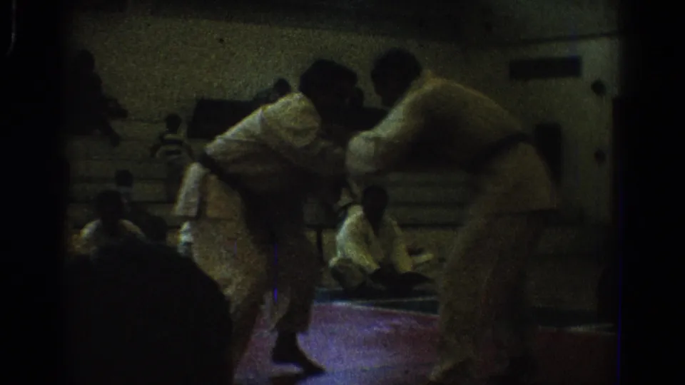 1966 West Covina 8mm frame of two uniformed men grappling low on a red indoor mat while people sit along the wall.