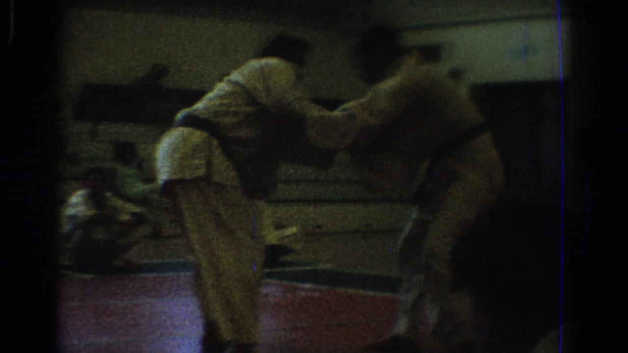 In 1966 West Covina home-movie footage, two men in white karate uniforms and black belts grapple on a red mat as spectators sit behind them.