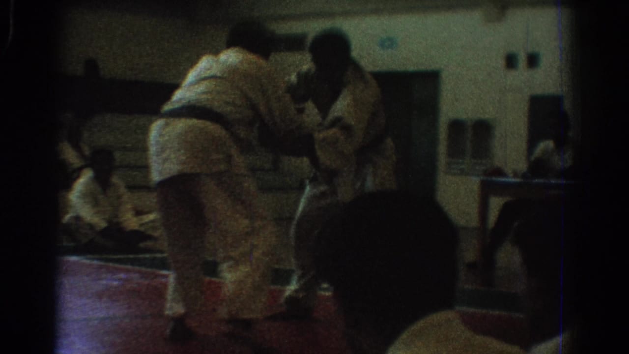 Karate competitors stand close together on a red mat with spectators nearby.