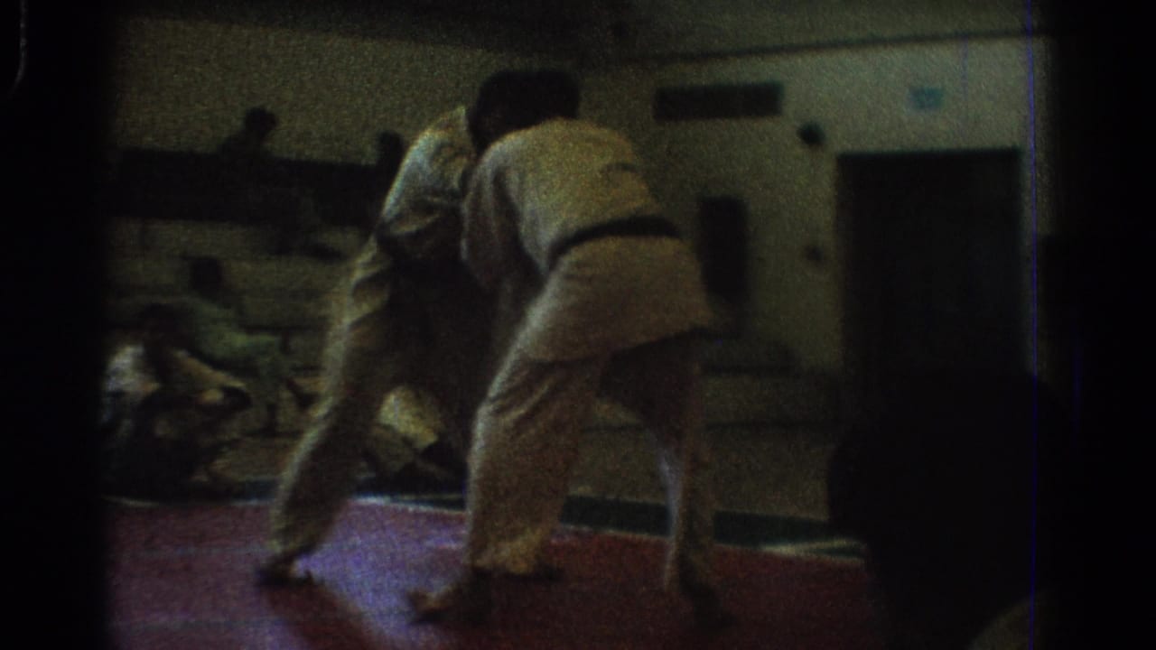 A dim 8mm frame shows one competitor bending forward as the other shifts behind him during the karate match on a red mat.