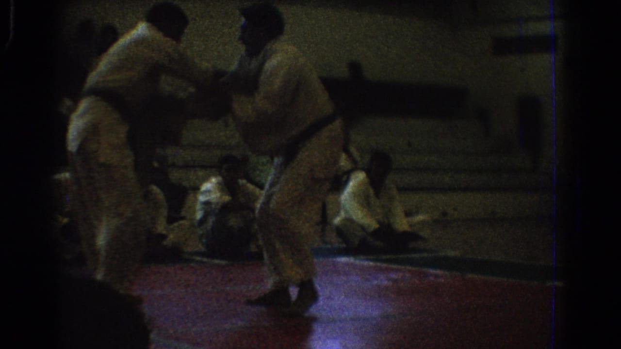 The two men face each other in white karate uniforms on the red mat, with spectators seated along the gym wall in the background.
