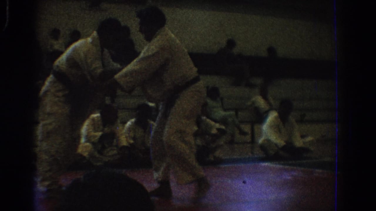 Two martial artists in white gi grapple on a red mat as spectators sit behind them.