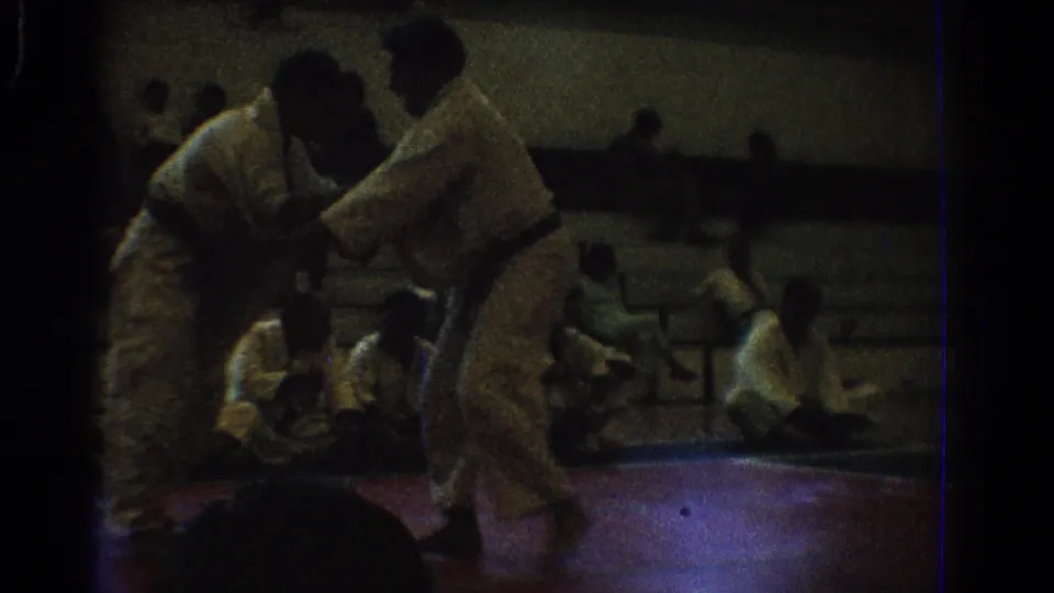 1966 West Covina home-movie frame of two martial artists in white gi grappling on a red mat inside a dim gymnasium.
